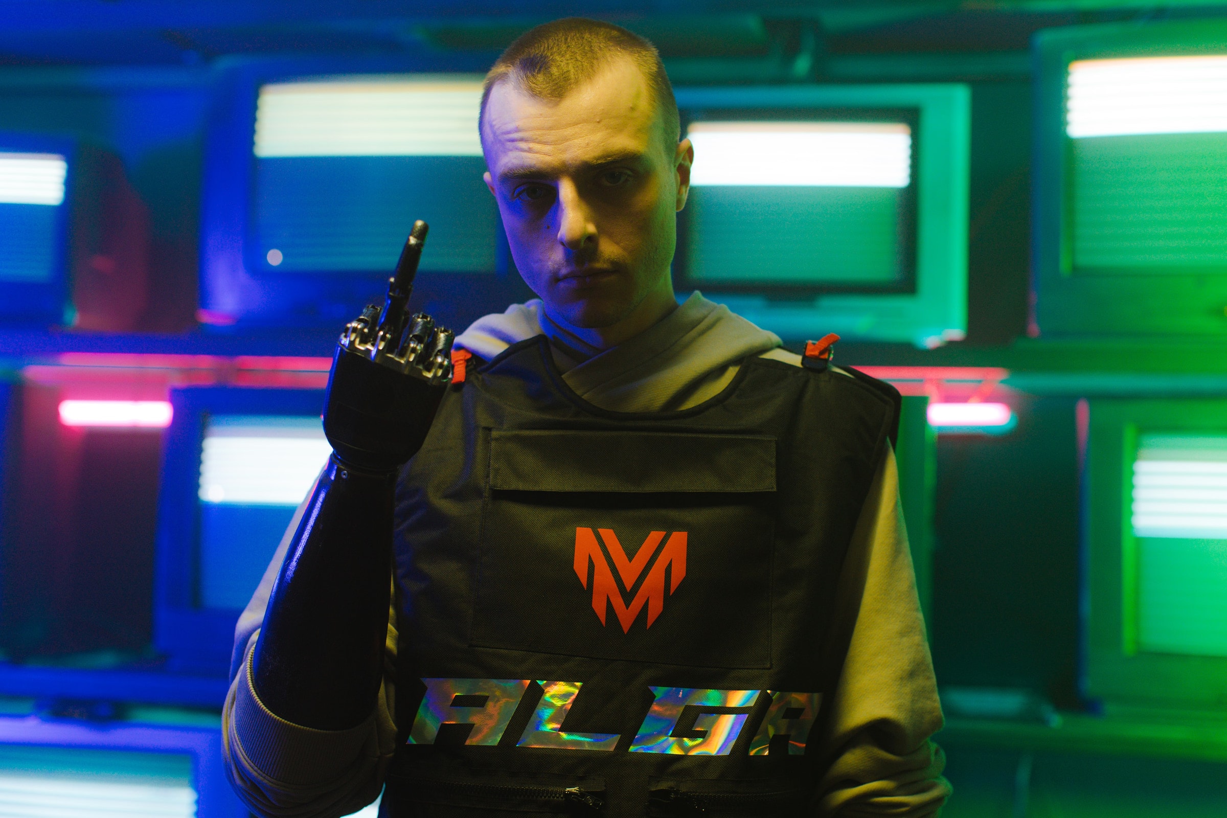 A man with a bionic arm stands confidently in a neon-lit room full of vintage TVs.