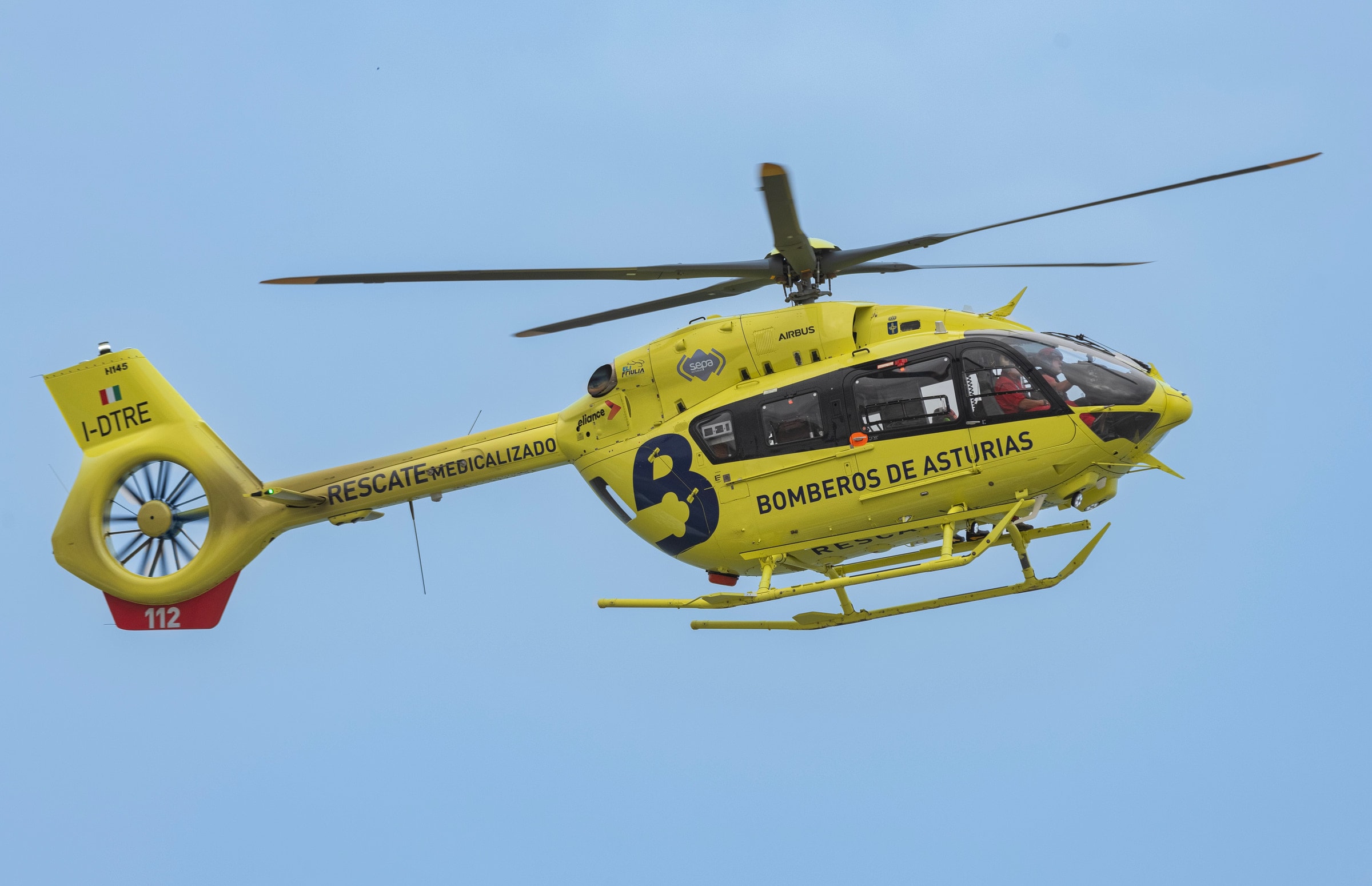 A yellow rescue helicopter from Asturias in flight with clear branding and emergency identification.