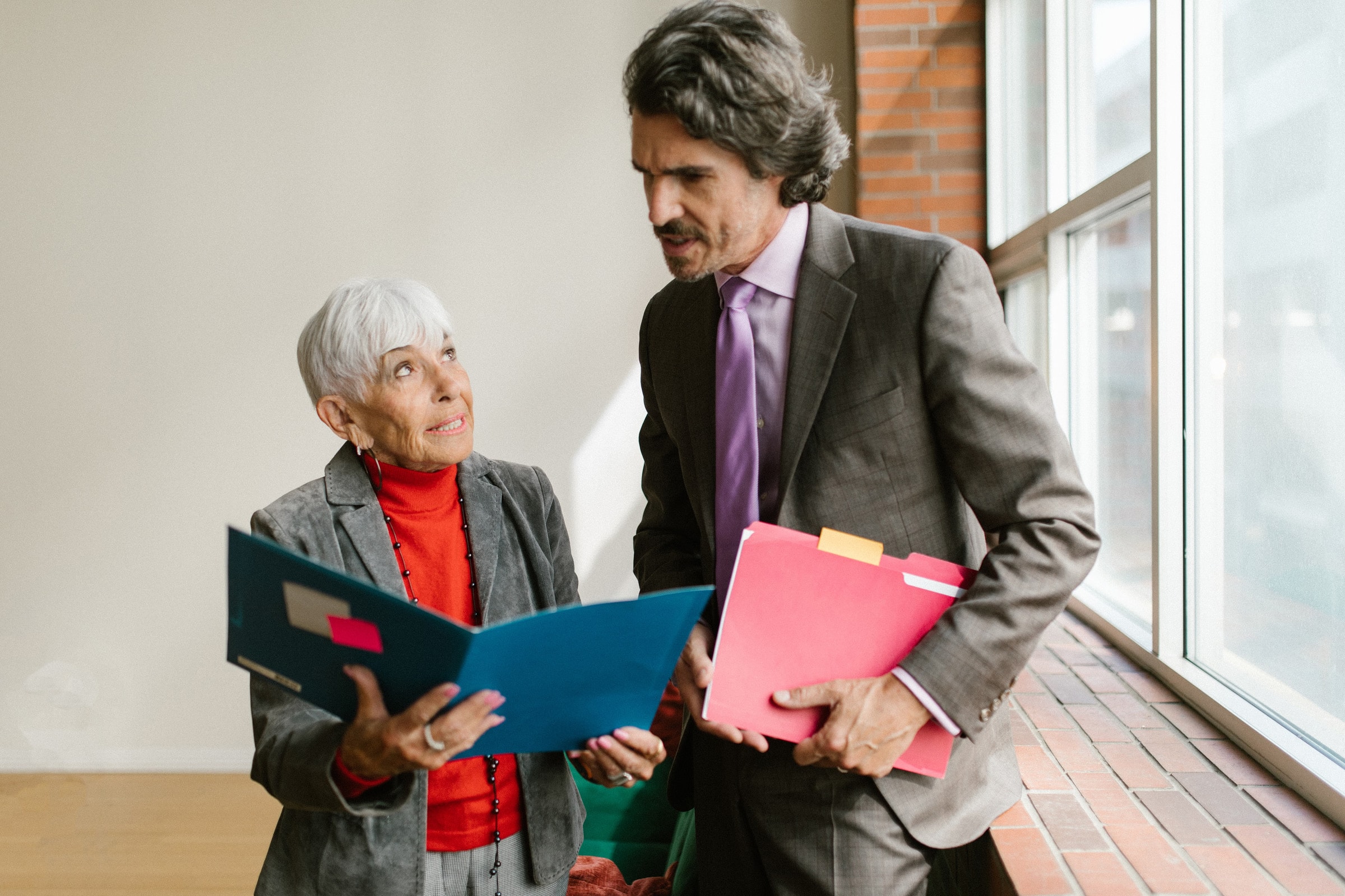Two senior professionals talking and reviewing business documents near an office window.